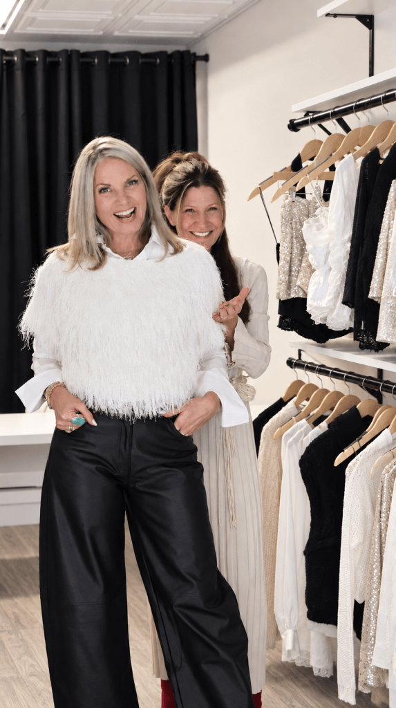 Two women smiling in a fashion boutique, one wearing a fluffy white top and black leather pants, while the other stands behind her in a light-colored dress.